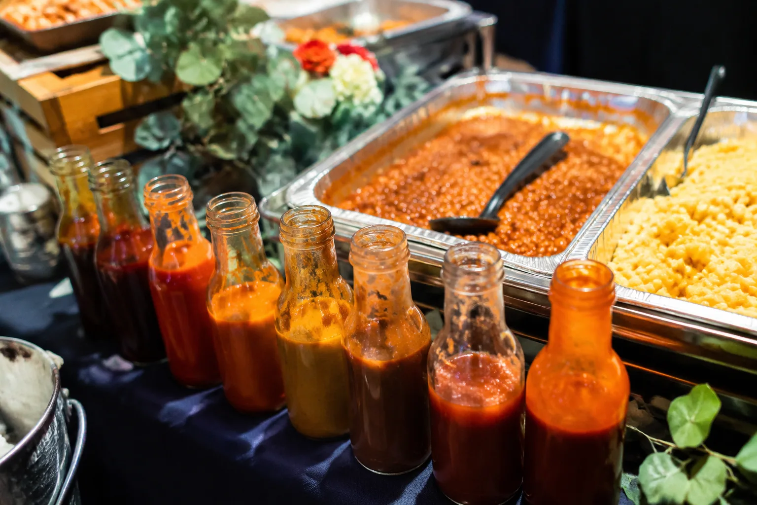 Small glass bottles of different barbecue sauces lined up on a catering buffet table next to labeled sides like baked beans and mac and