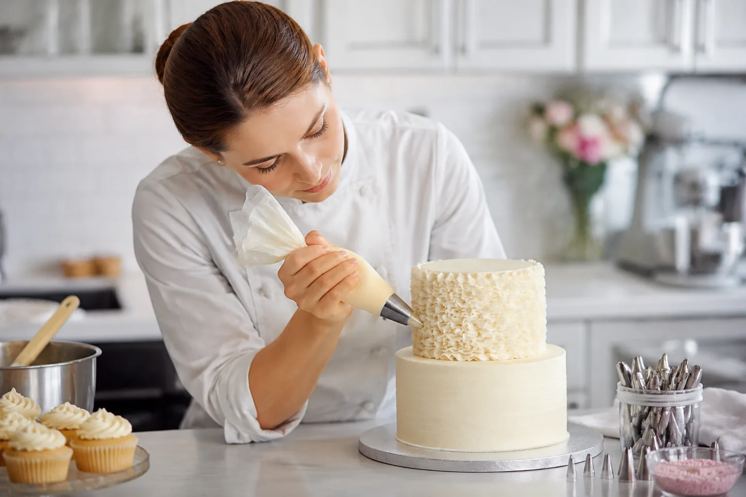 Baker piping delicate buttercream texture onto a tiered cake in a bright, clean bakery kitchen, decorating tools visible on the counter