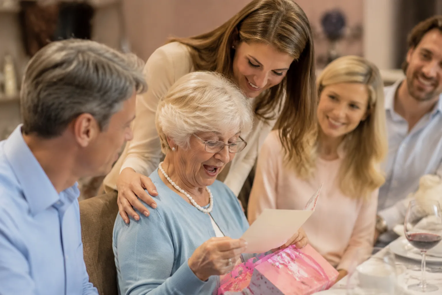 A photorealistic image of a grandmother reading a birthday card handed to her by her adult grandchild at a family dinner table, warm candid