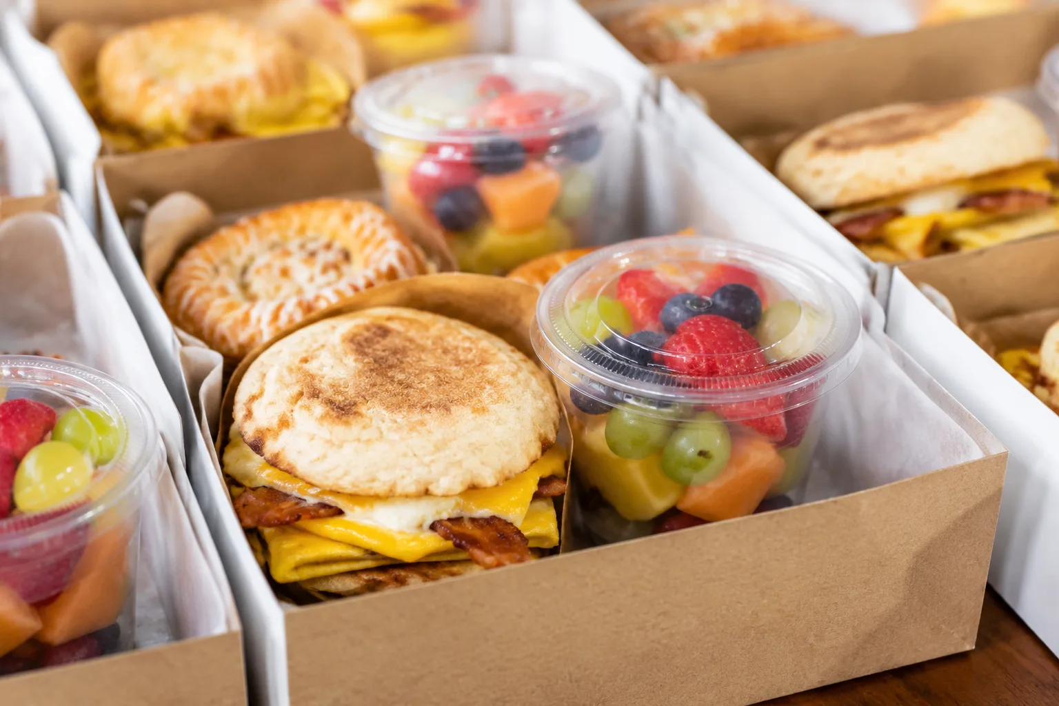 A photorealistic close-up of individually packaged breakfast boxes with a breakfast sandwich, fruit cup, and pastry, arranged for a catered