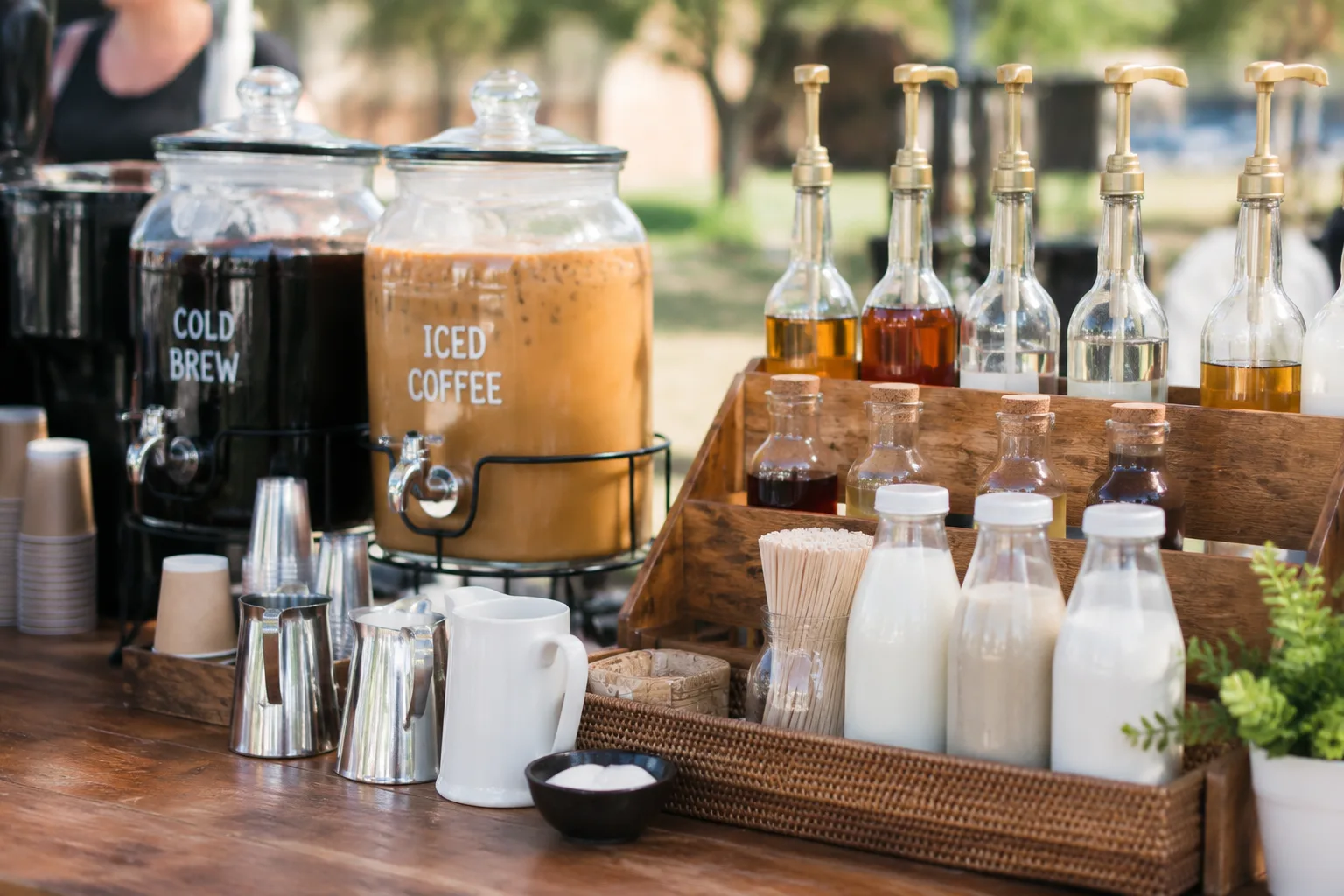 A photorealistic close-up of a self-serve coffee station with iced coffee, cold brew, and a variety of milk and syrup options at an outdoor