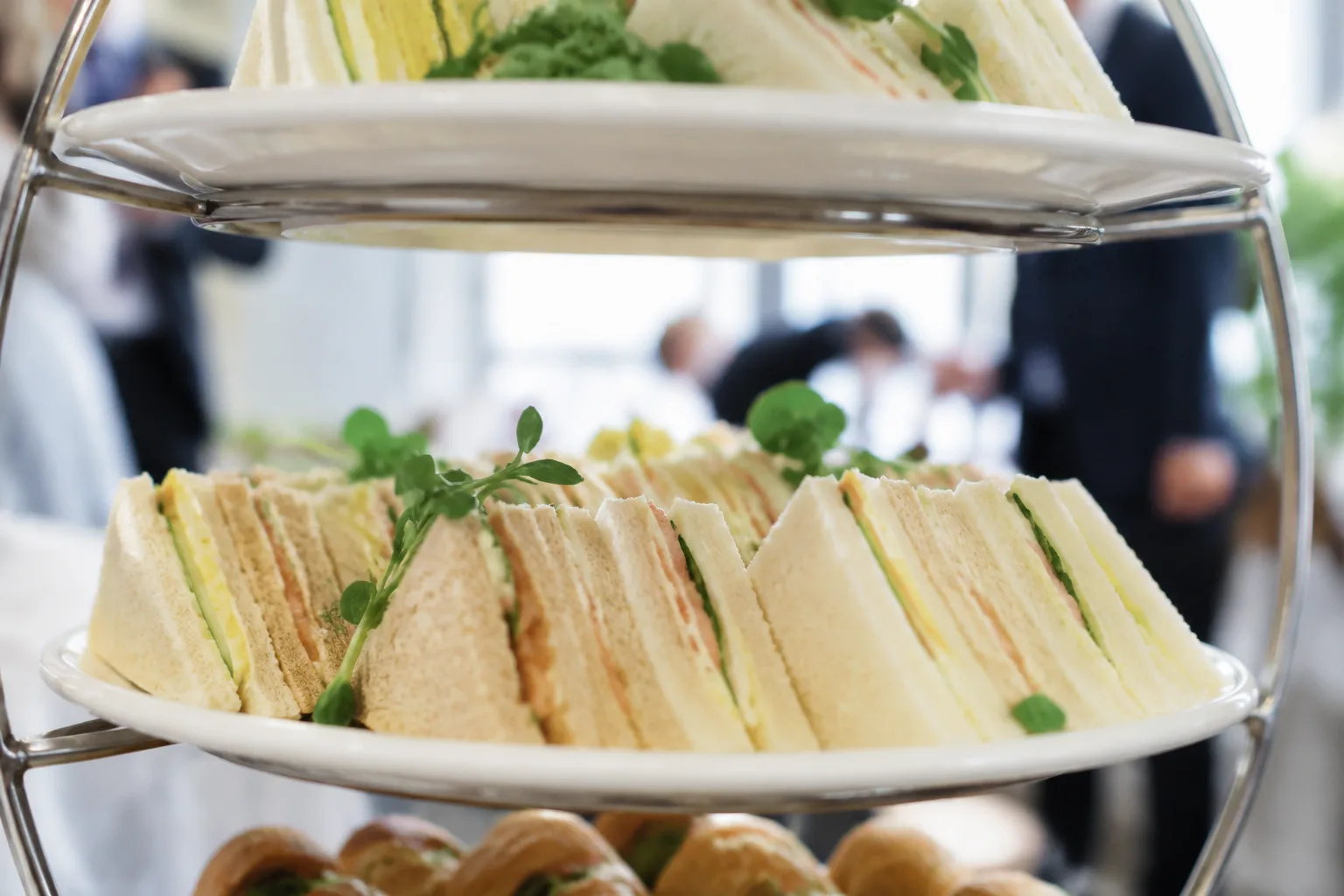 A photorealistic close-up of small triangle-cut finger sandwiches arranged on a tiered serving tray at an office gathering