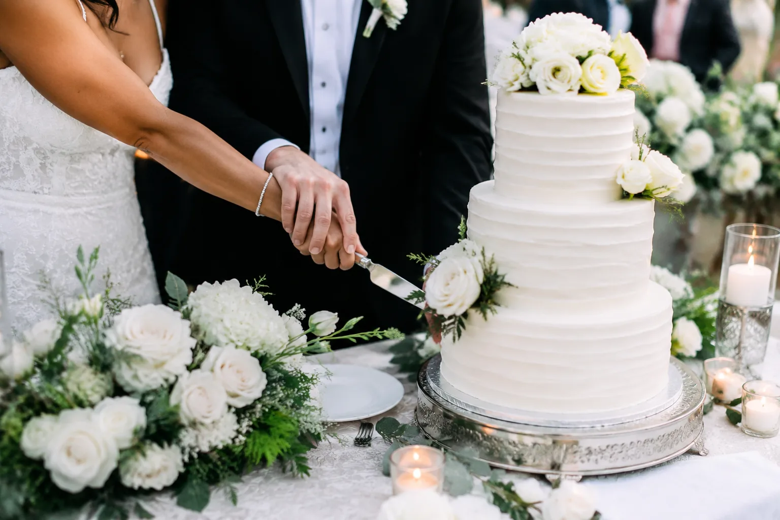Elegant multi-tier wedding cake being cut by a couple at a reception table decorated with white florals, close-up candid shot