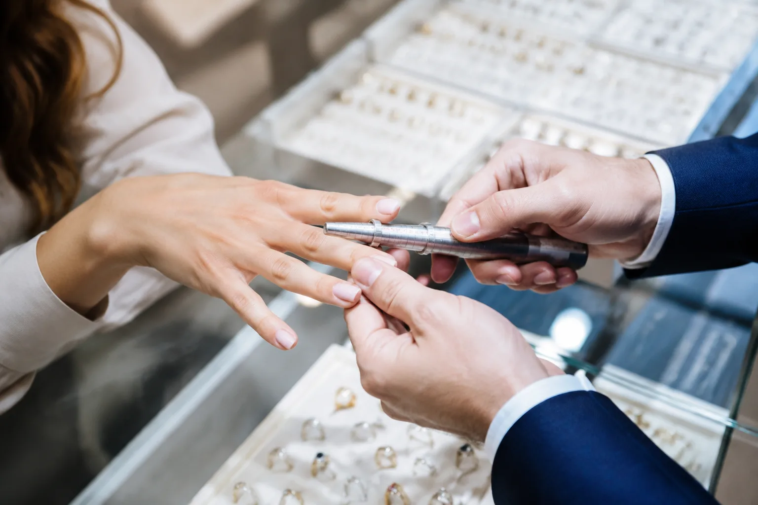 Jeweler's hands using a ring sizing tool to measure a customer's finger at a jewelry counter, bright clean lighting, ring trays visible in