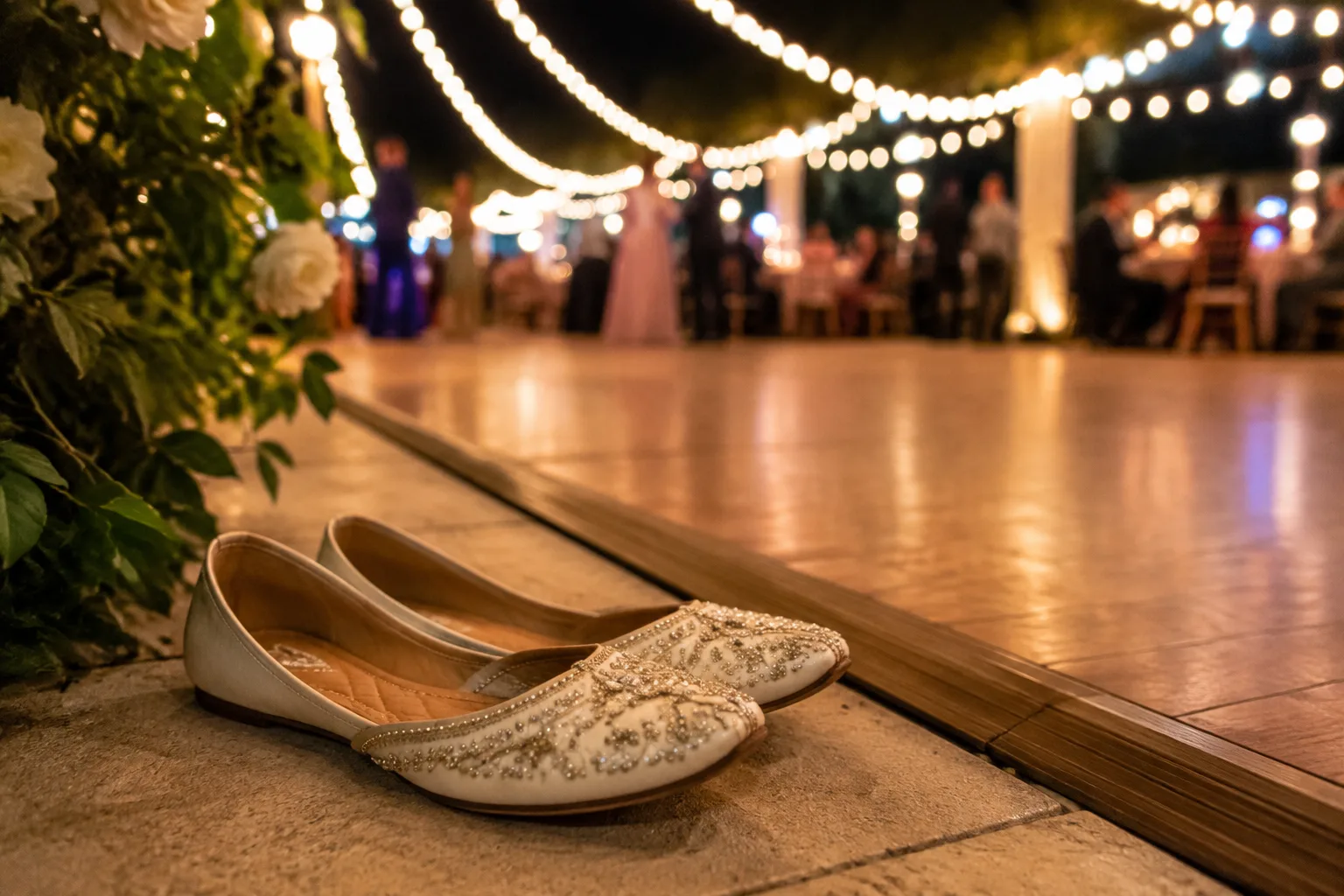Pair of embellished bridal flats placed neatly beside a dance floor at an evening wedding reception, ambient string